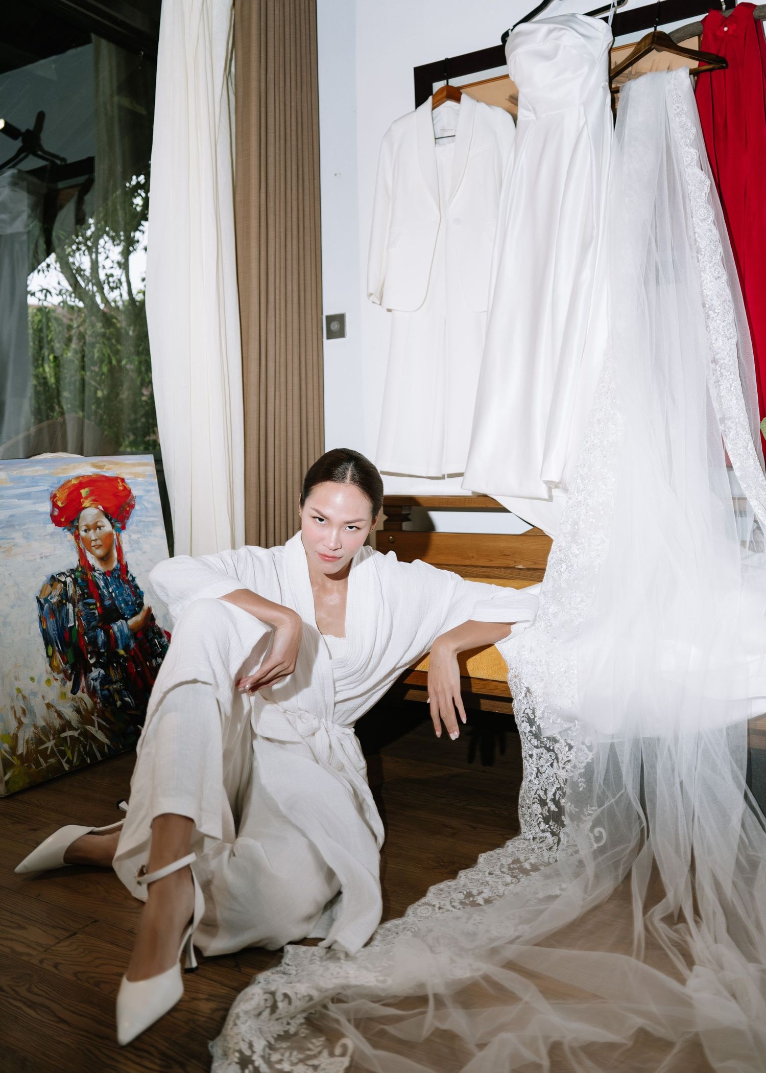 Bride in white robe sitting on floor near hanging wedding dresses and colorful portrait painting.