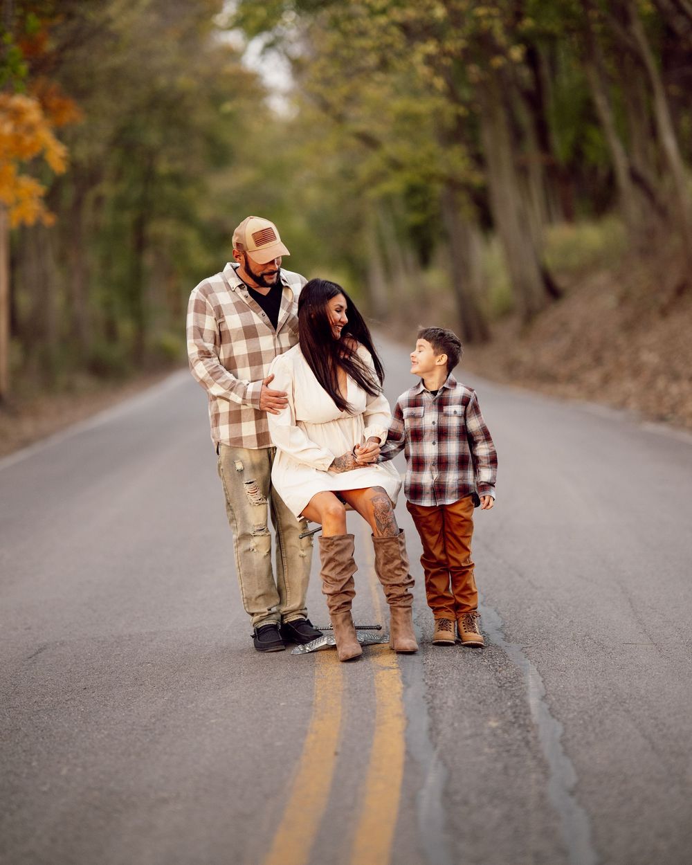 A family wearing matching plaid stands together on an empty autumn road surrounded by trees.