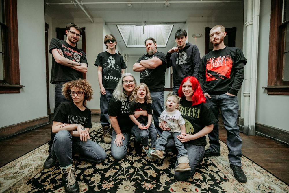 A group of people in casual clothing and band t-shirts pose together in a hallway with patterned carpet.