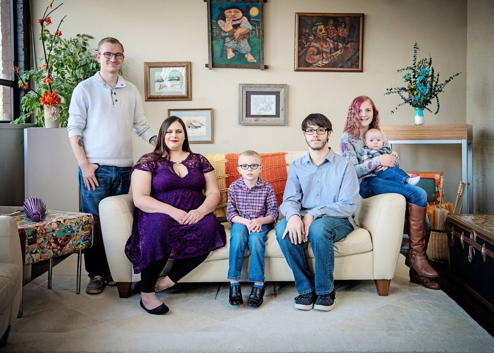 Group photo of five people sitting together on a light colored couch with wall art and plants in the background.