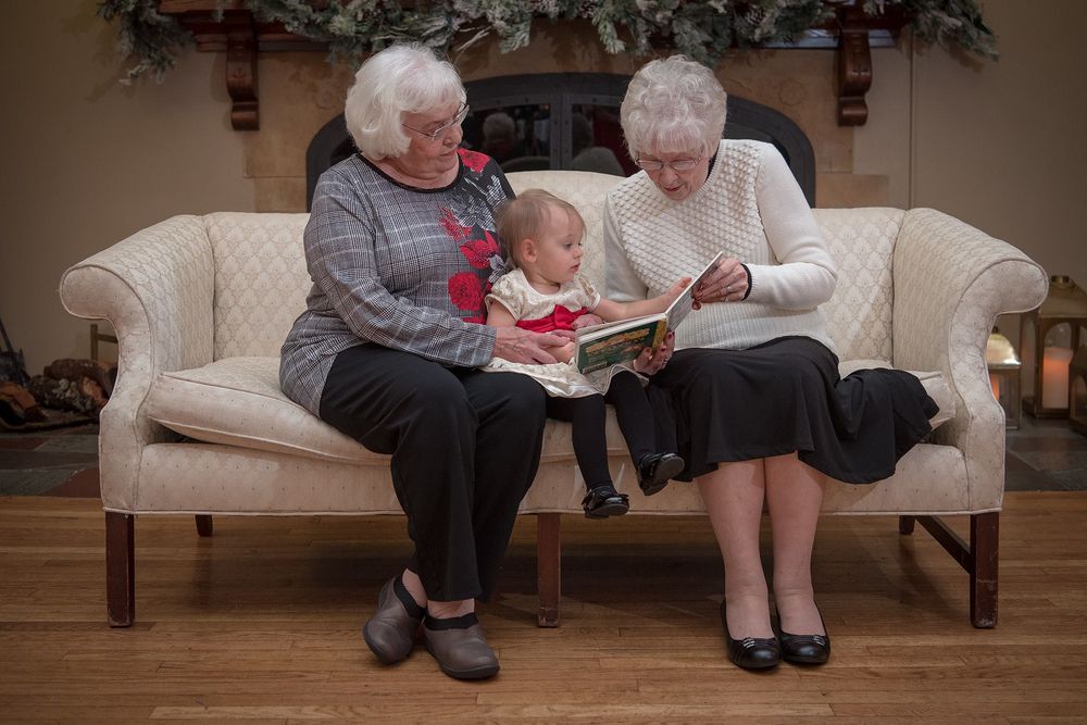 Two seniors reading a book with a small child on a beige couch.
