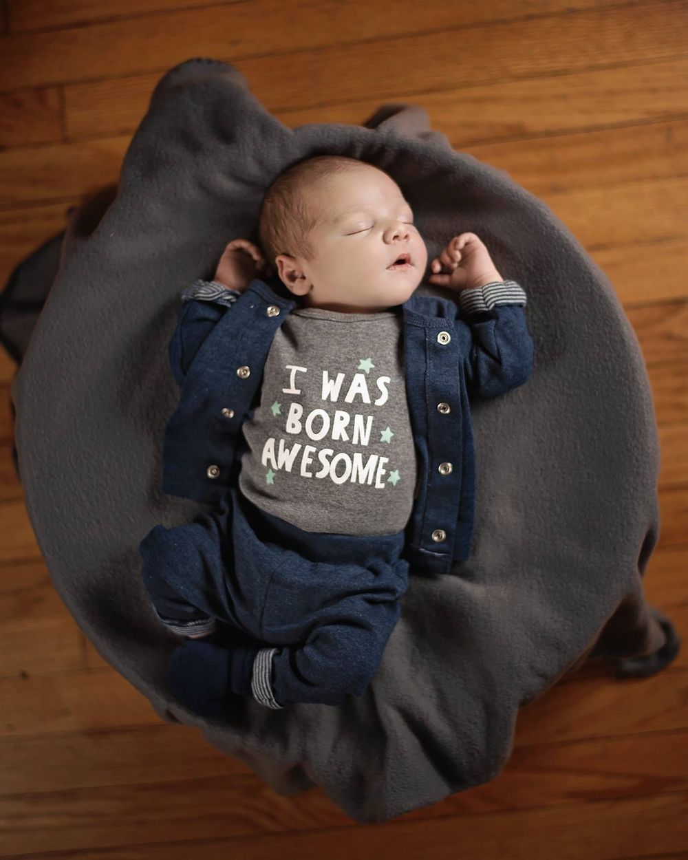 Newborn baby lying on grey blanket wearing navy outfit with text saying "I was born awesome".
