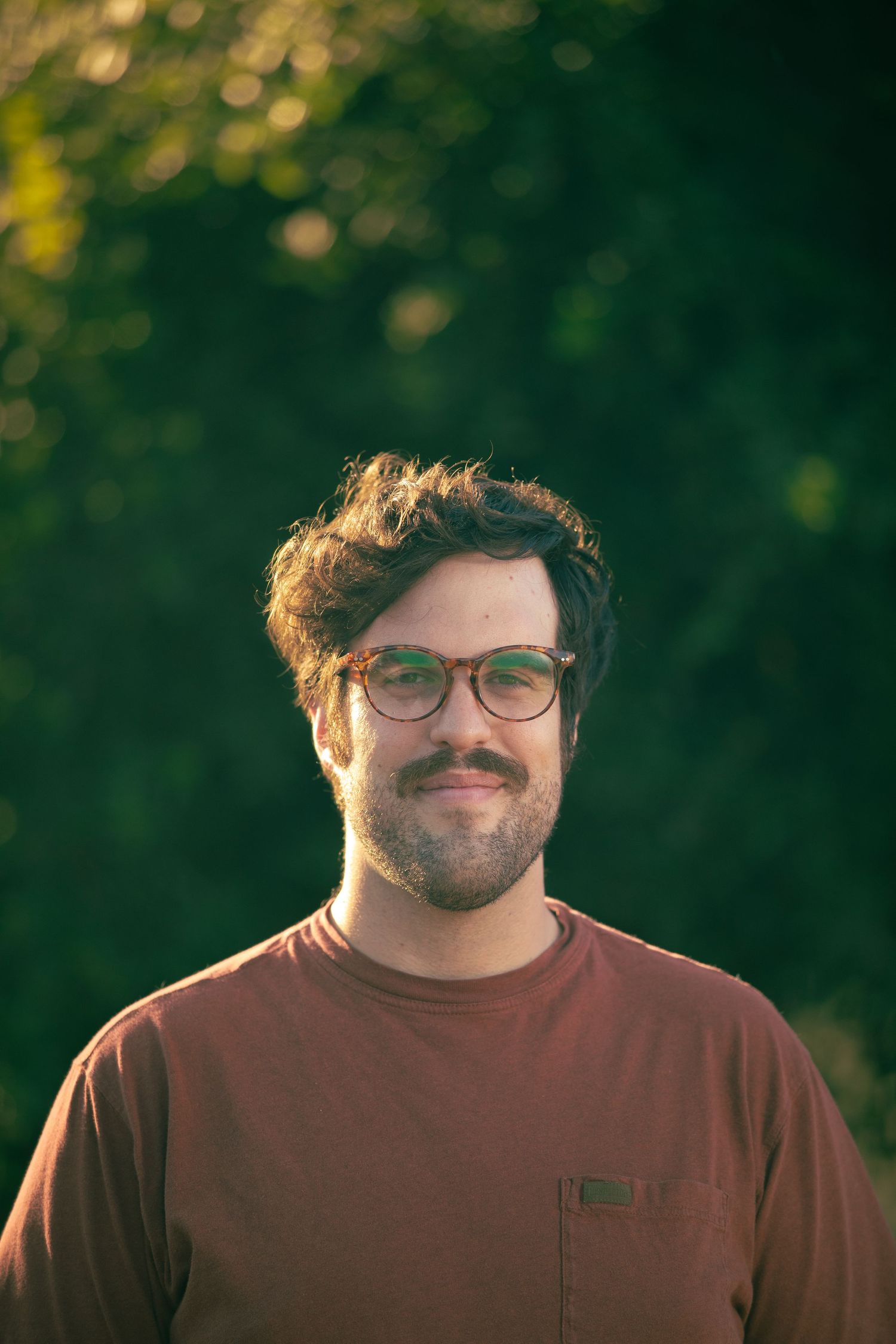 Person in burgundy shirt smiling warmly outdoors with sunlit trees in background.