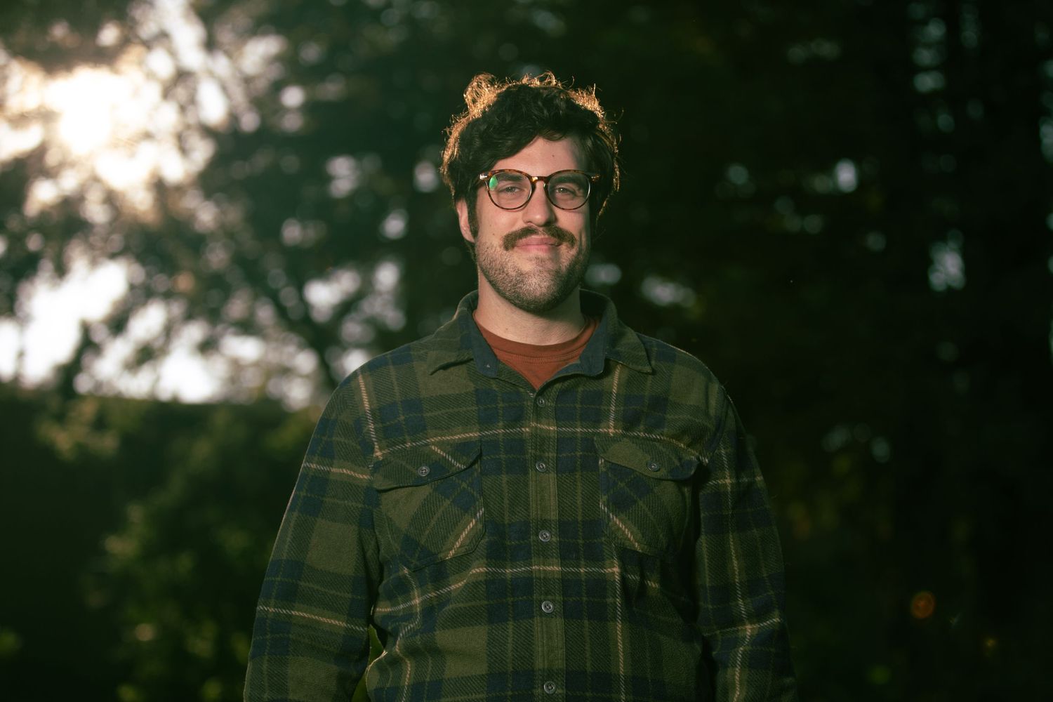 Portrait in warm evening light with a brown plaid shirt against blurred natural background.