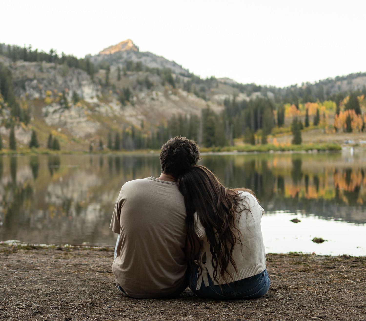 Couple sitting by Tony's Grove lakeshore during their couples session. Looking out towards the lake and beautiful forest and mountain peaks.