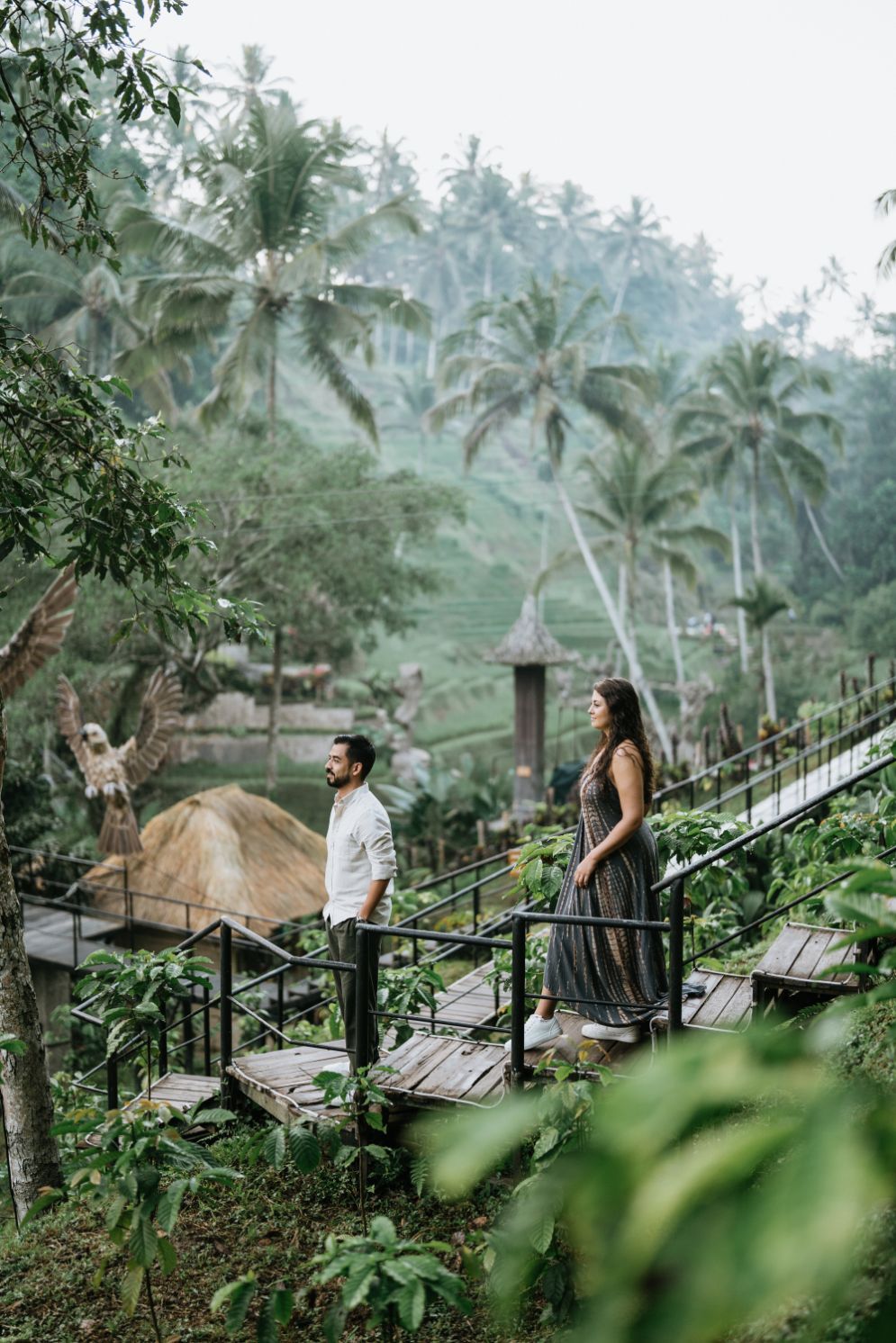 Two people walk along an elevated wooden pathway through a tropical palm forest.
