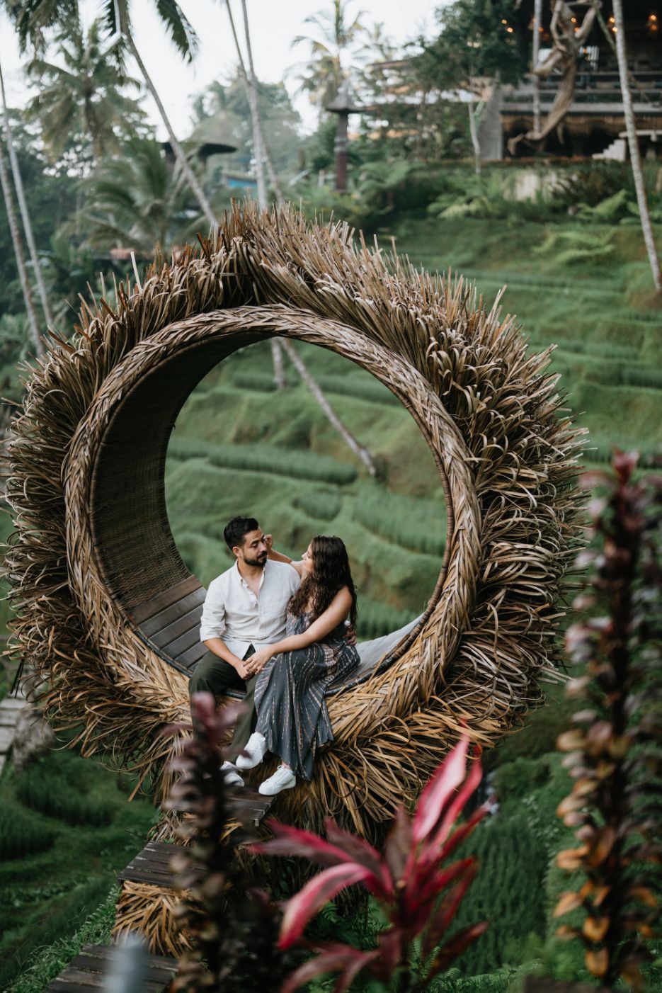Couple sits in a large woven nest-like swing overlooking tropical rice terraces.