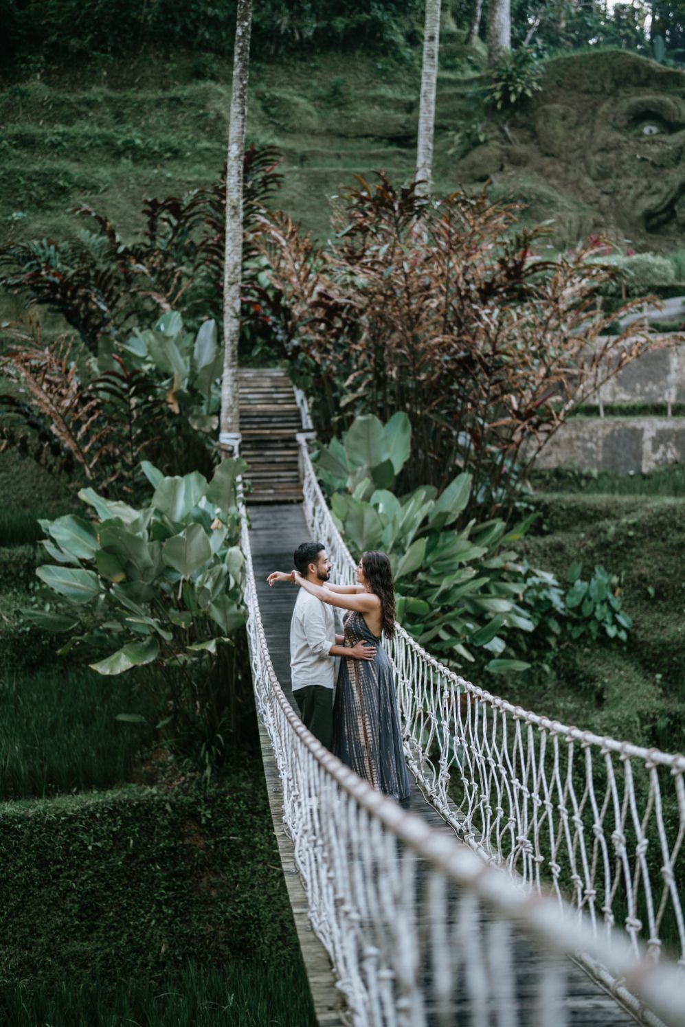 Couple shares an intimate moment on a rope bridge surrounded by tropical foliage.
