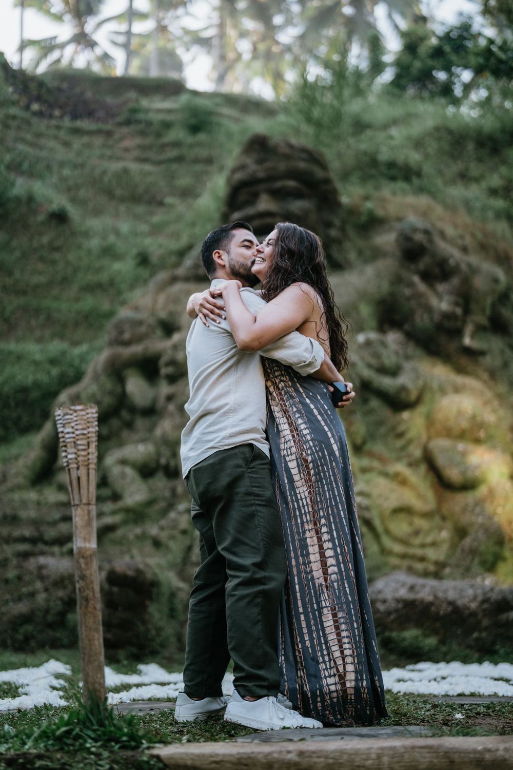 A couple embraces lovingly on a wooden bench surrounded by tropical landscaping and stone features.
