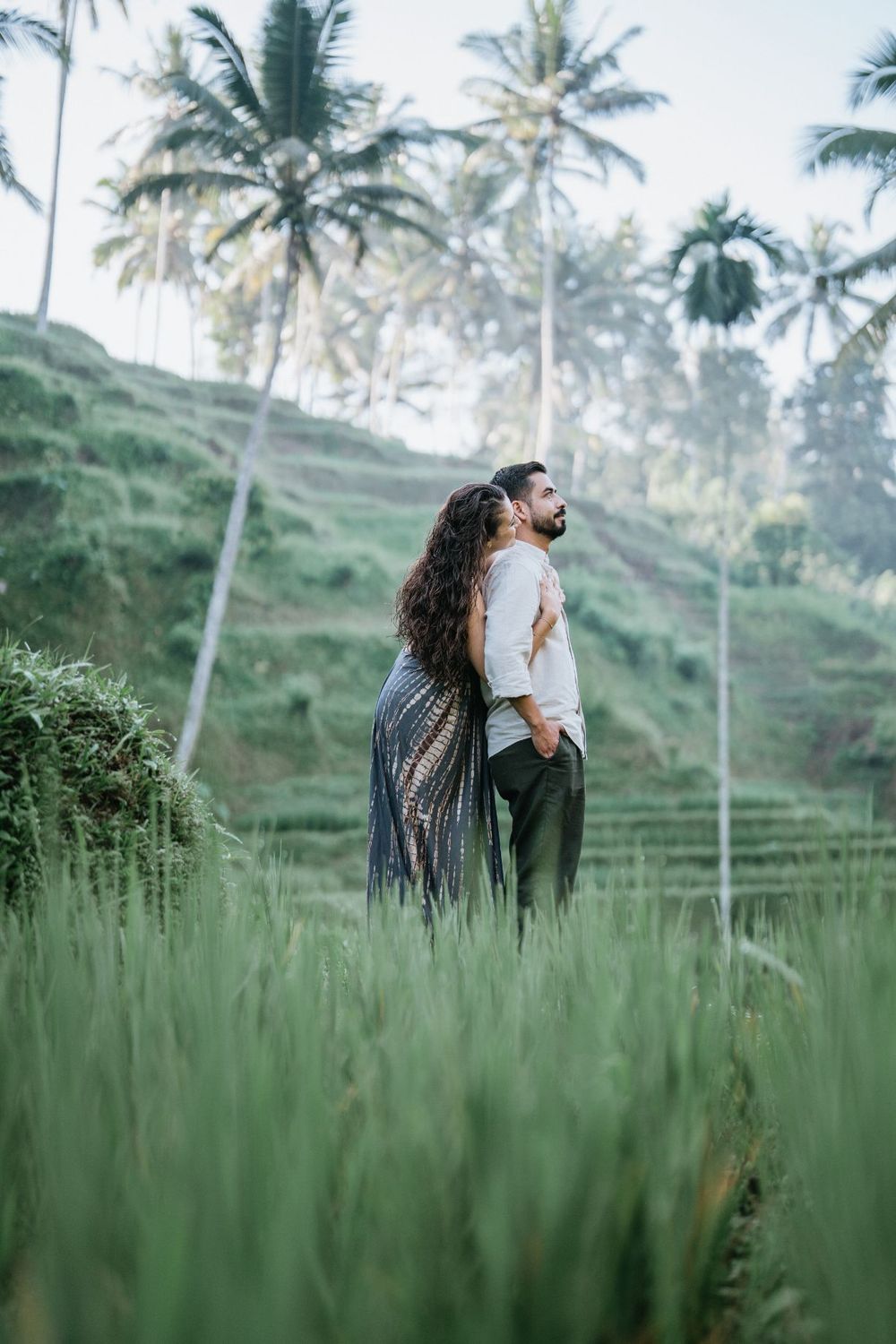 A romantic couple stands among tall grass with palm trees in the background during golden hour.