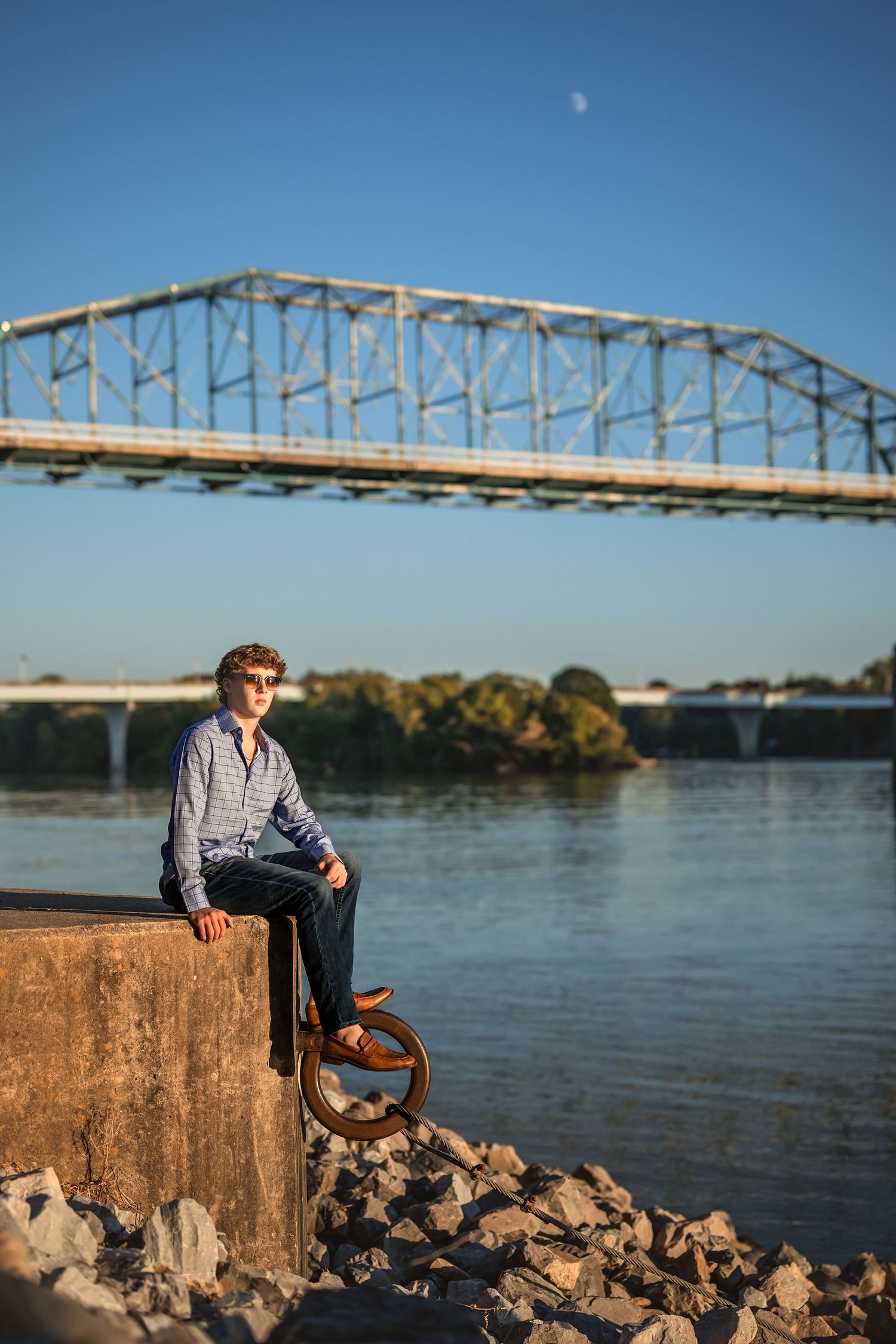 Senior boy sits on a concrete ledge near the Tennessee river with the Market St walking bridge and moon visible in the blue sky.
