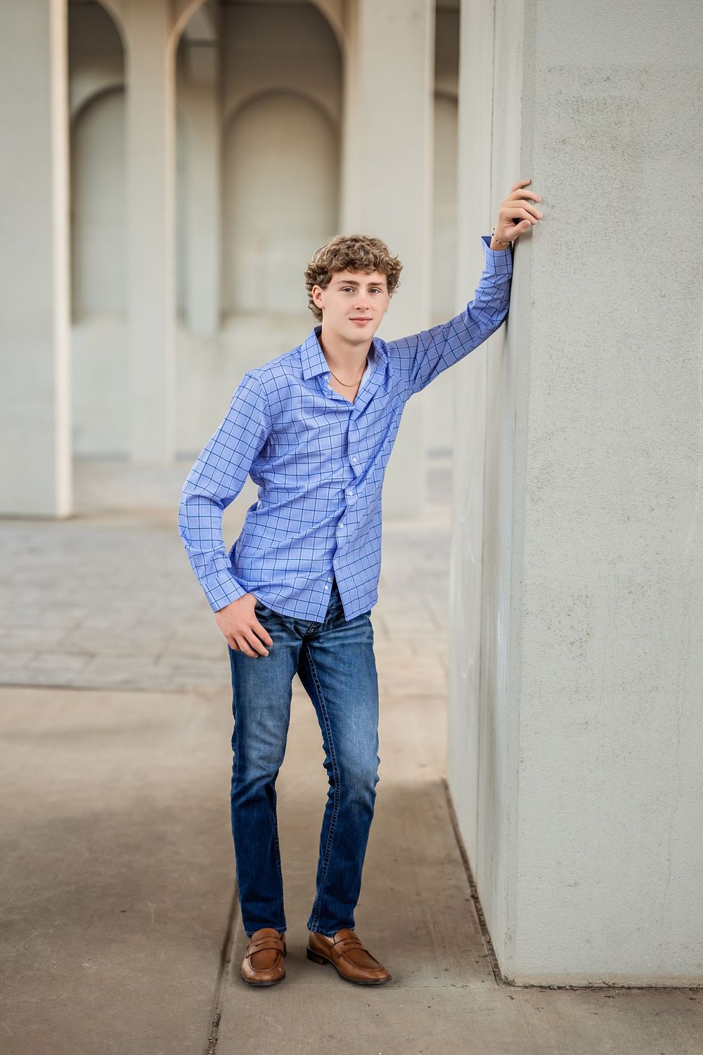Senior boy wearing a blue plaid shirt and jeans poses dynamically under concrete archways in an editorial style.