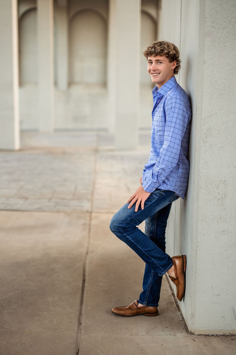 A senior boy in a blue plaid shirt and jeans poses casually against a white outdoor column under the John Ross bridge in Coolidge Park.