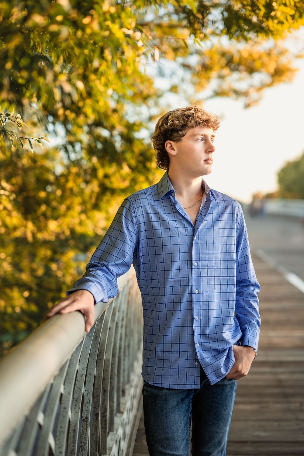 A senior boy in blue stands on the Chattanooga walking bridge in Autumn with golden foliage in the background.