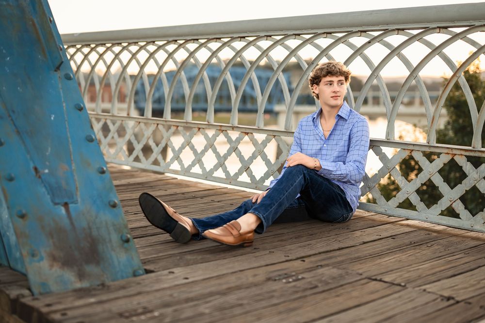 A senior boy sits on the Market St walking bridge over the Tennessee river during golden hour.