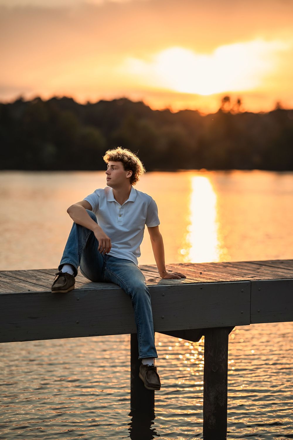 A senior boy lounges on a wooden pier at sunset with the orange sky reflecting off the calm water.