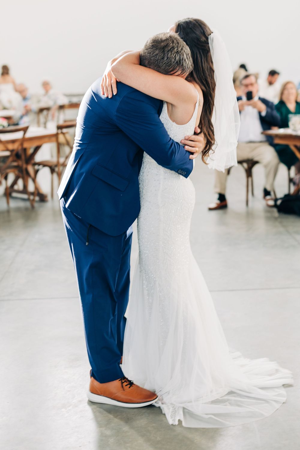 A bride and groom share an intimate first dance in a bright indoor venue with guests seated in the background.