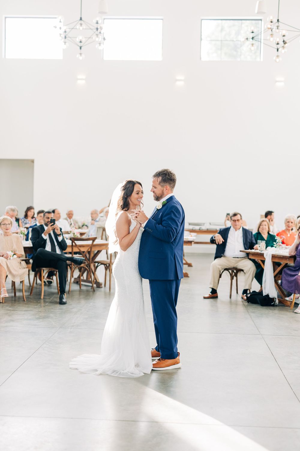 A couple shares their first dance in a bright reception hall while wedding guests look on from rustic wooden chairs.