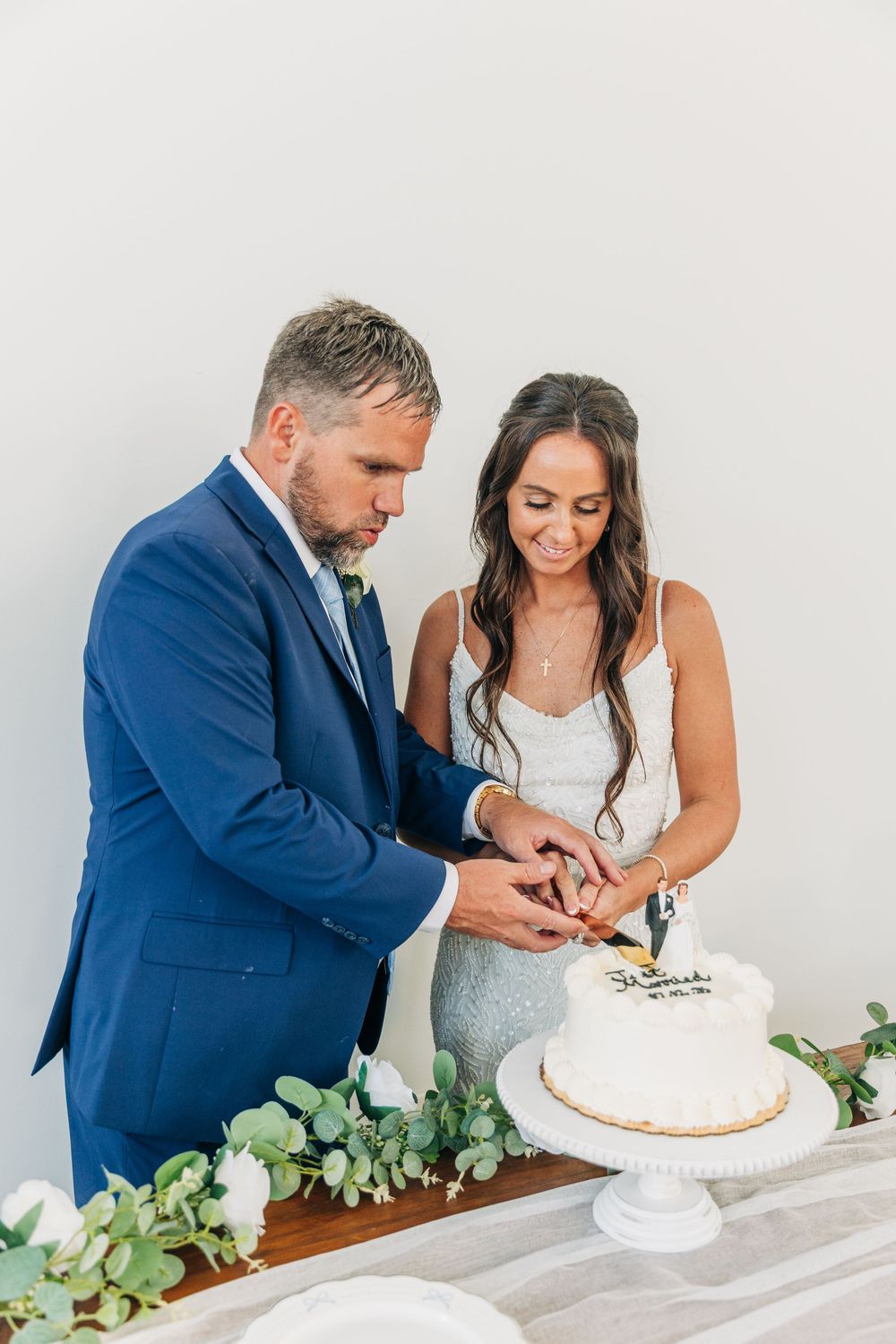 Newlyweds cut their elegant white wedding cake together on a pedestal decorated with eucalyptus greenery.
