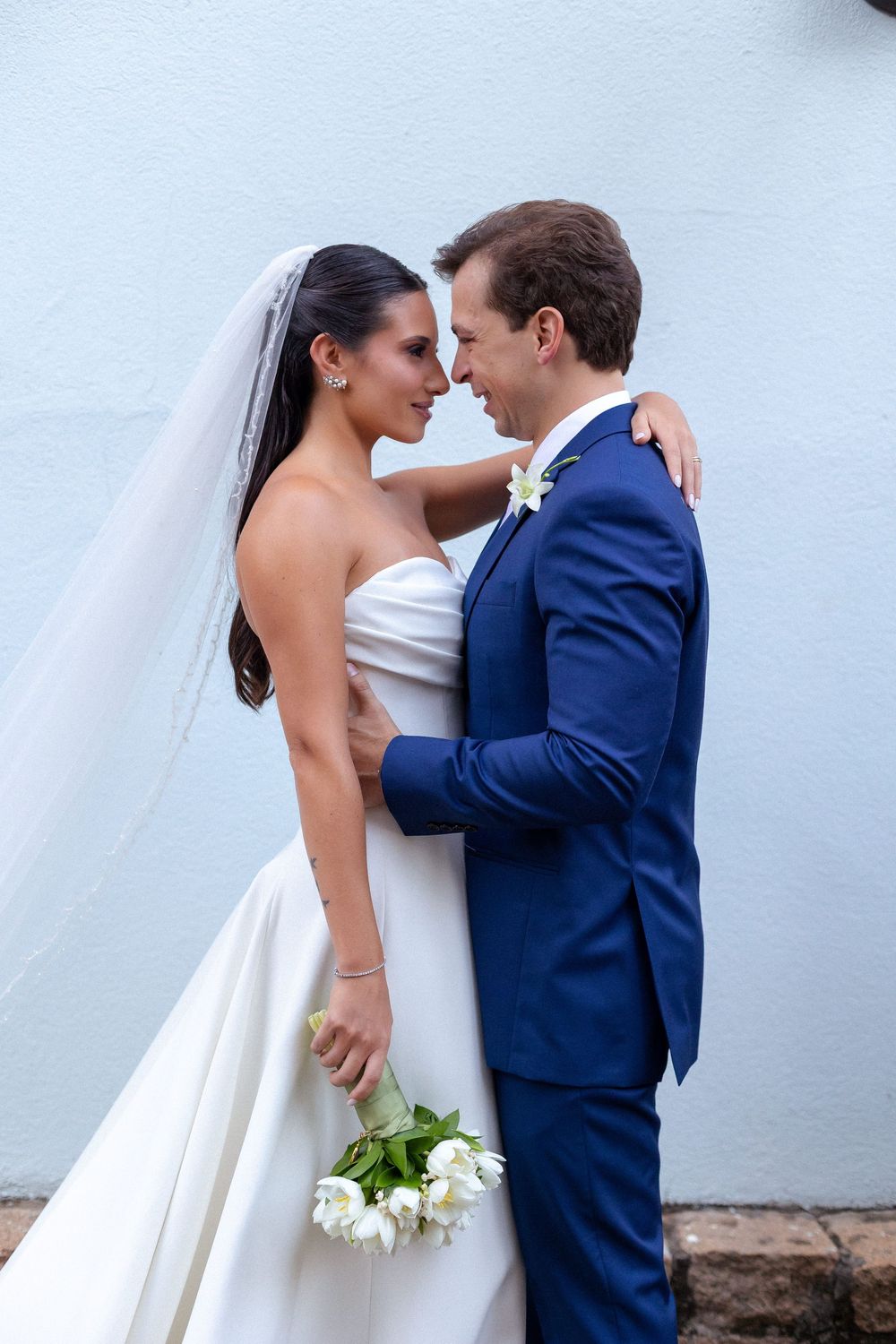 Bride in white gown holds white bouquet while embracing groom in navy suit against light wall.