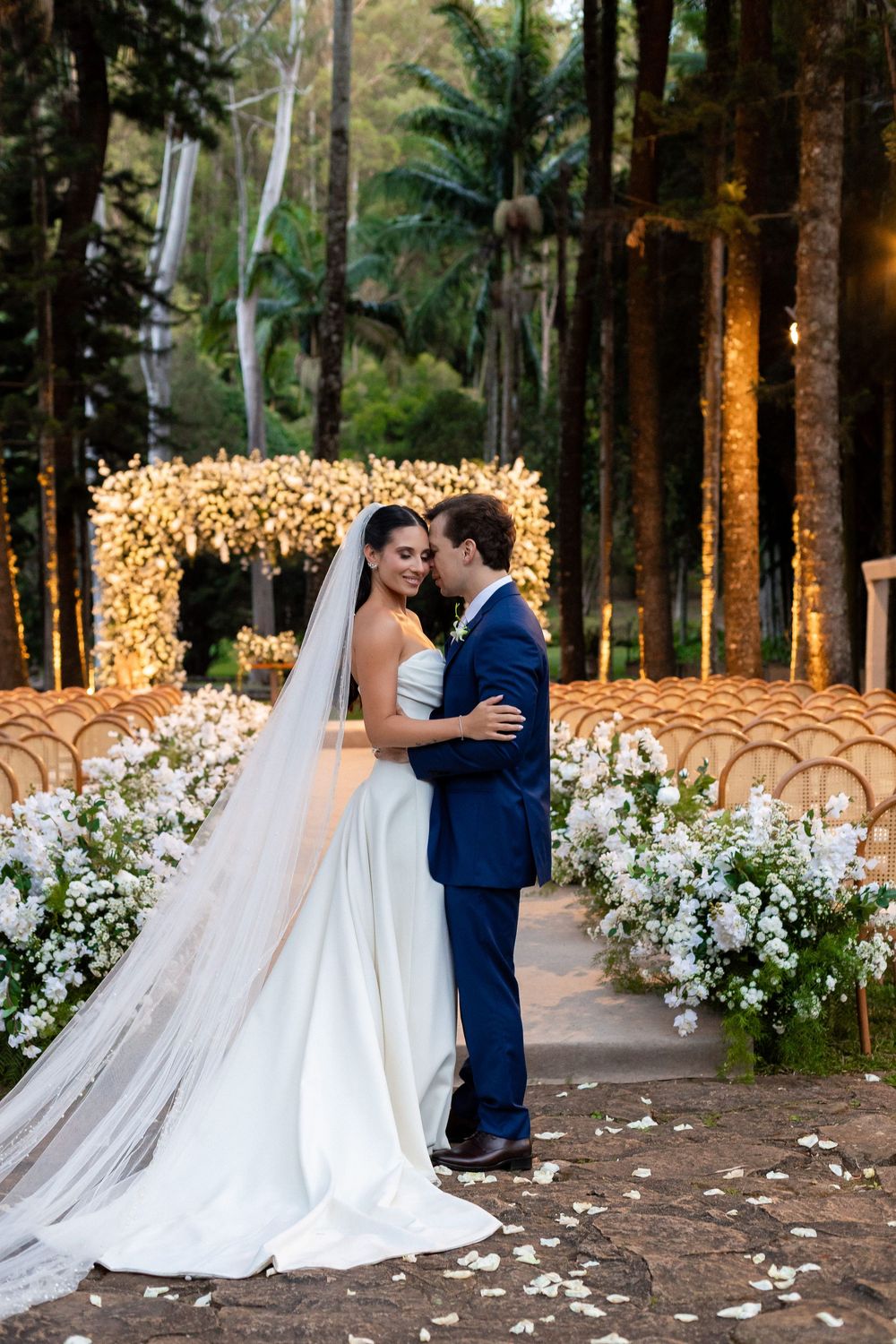 Romantic wedding ceremony in a forest setting with white floral decorations and glowing fairy lights in the background.