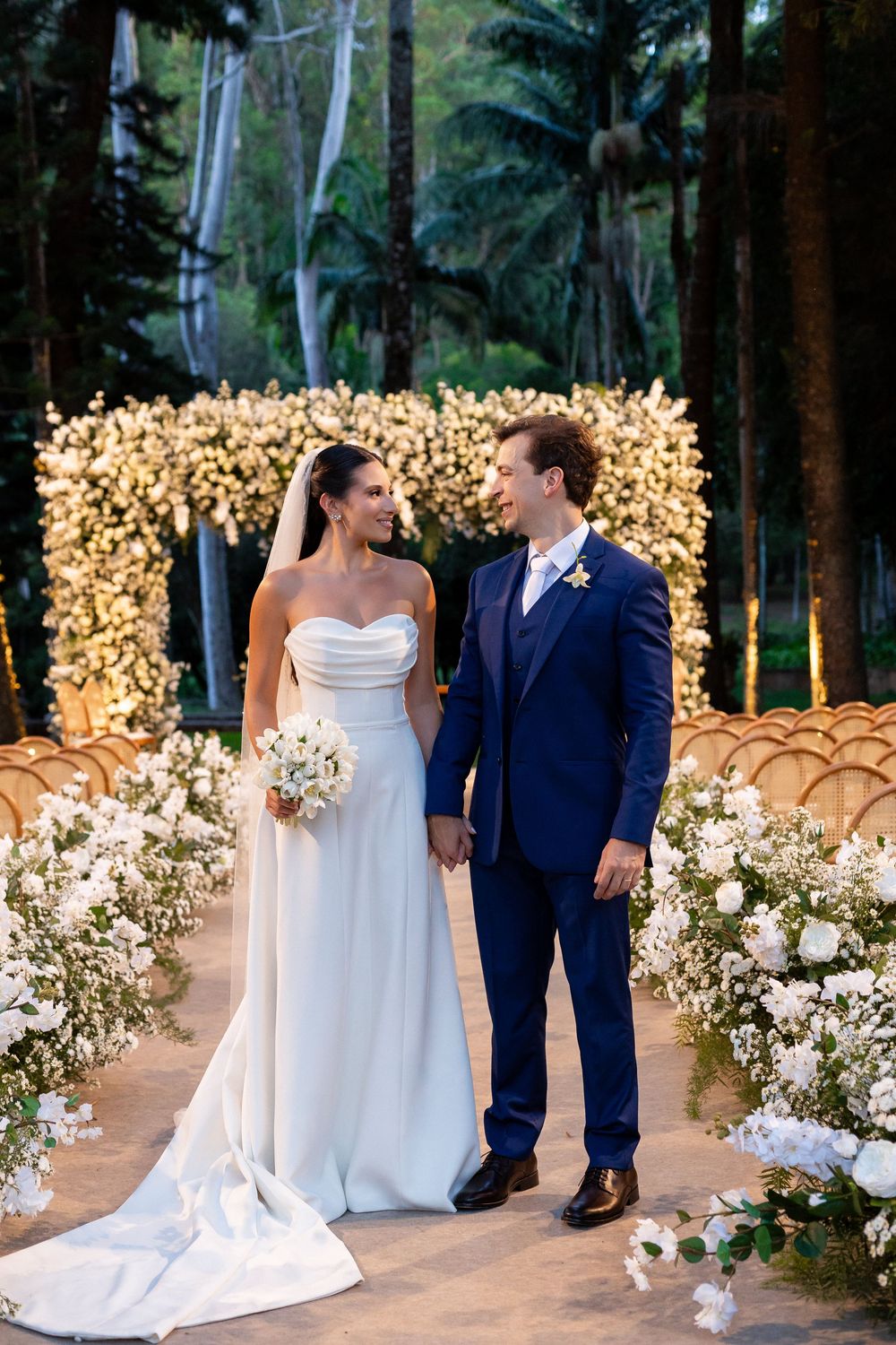 Elegant outdoor wedding ceremony setup with glowing light backdrop and white floral arrangements lining the aisle.
