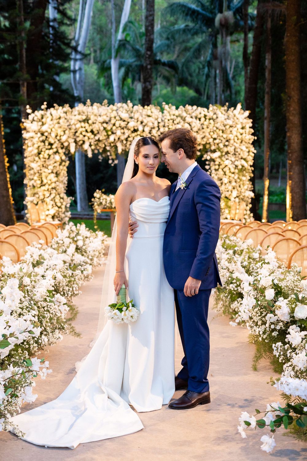Elegant outdoor wedding ceremony setup with white flower arrangements and golden light filtering through forest trees.