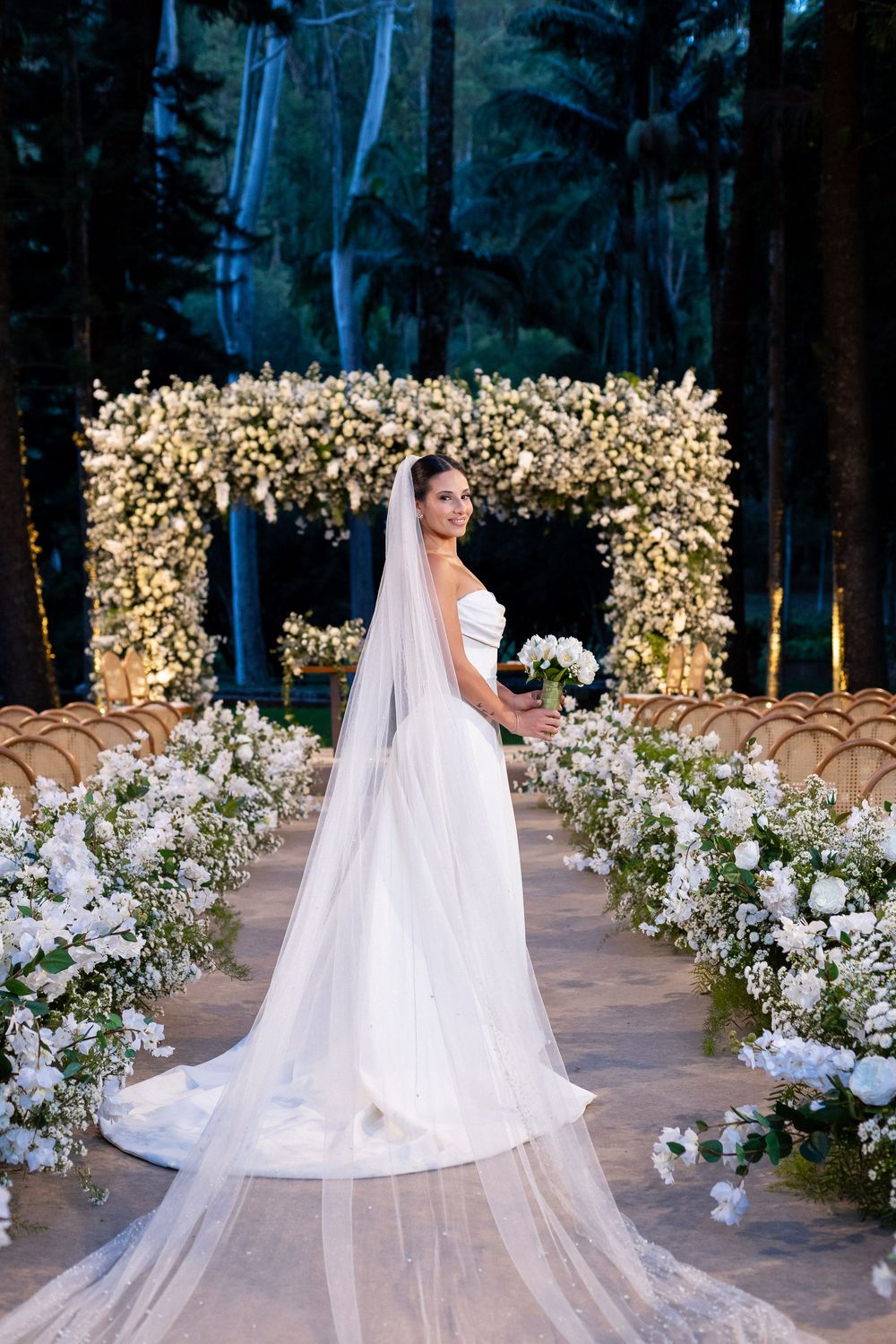 Elegant outdoor wedding setup with white floral arrangements and glowing lights along a stone pathway.