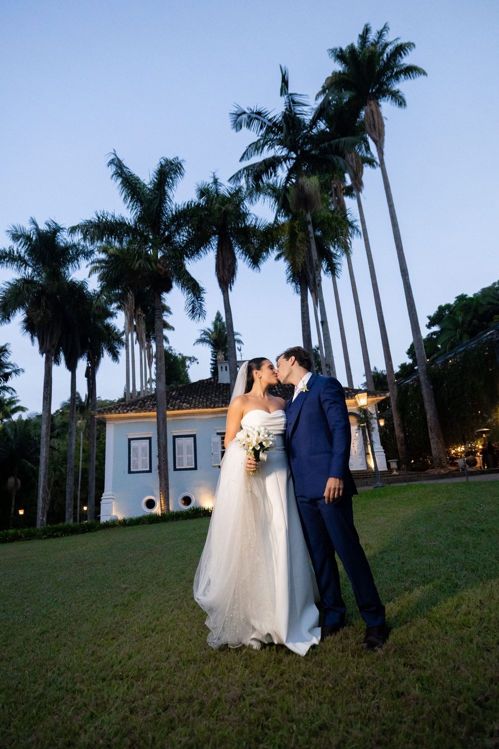 Newlyweds share romantic moment under towering palm trees at twilight beside historic white building.