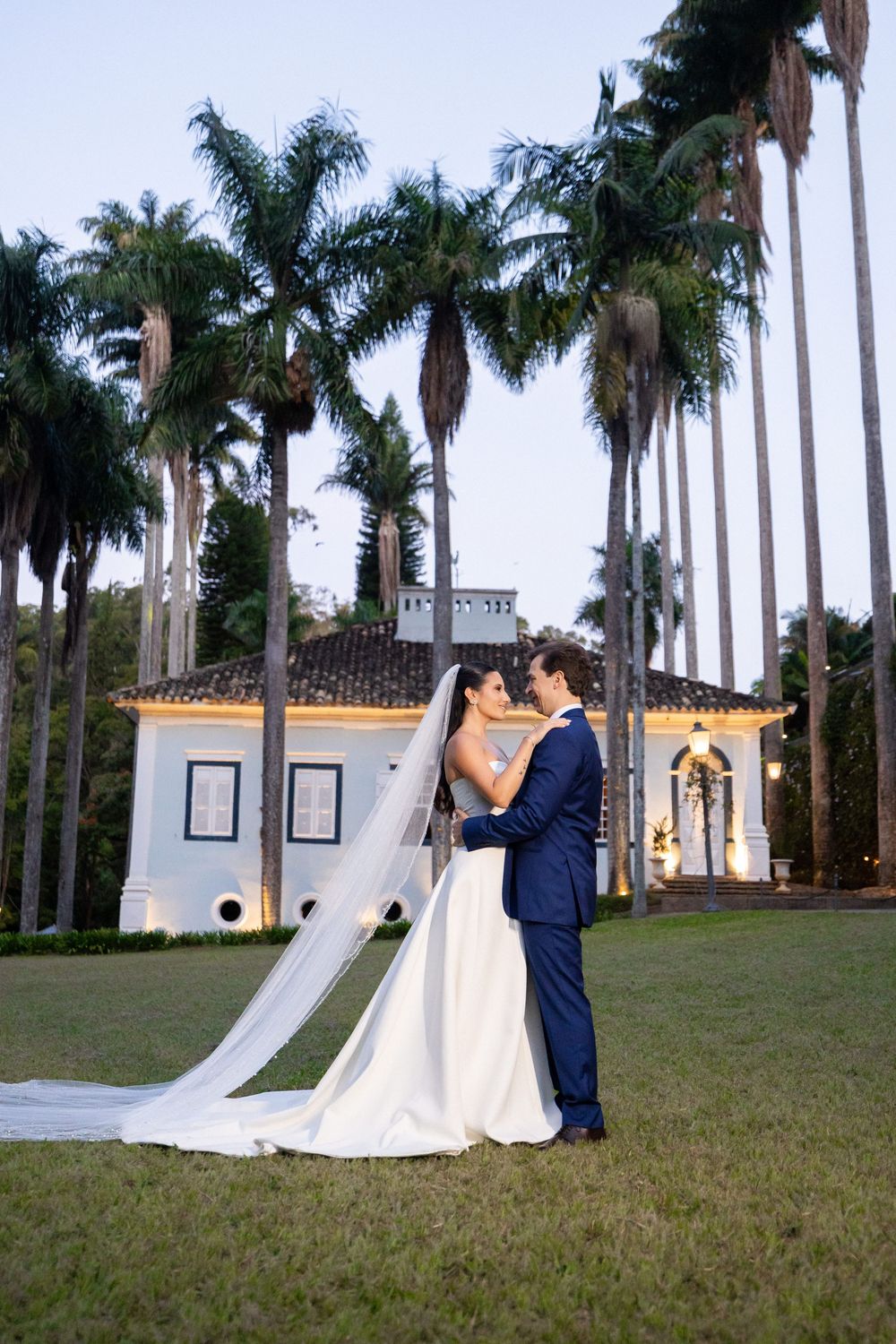 A couple poses together on a lawn surrounded by tall palm trees and a grand white building in the background.