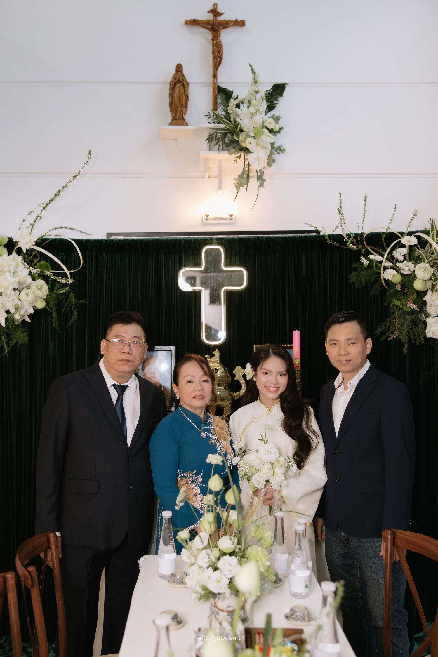 Wedding party poses before illuminated cross altar with white floral decorations in church setting.