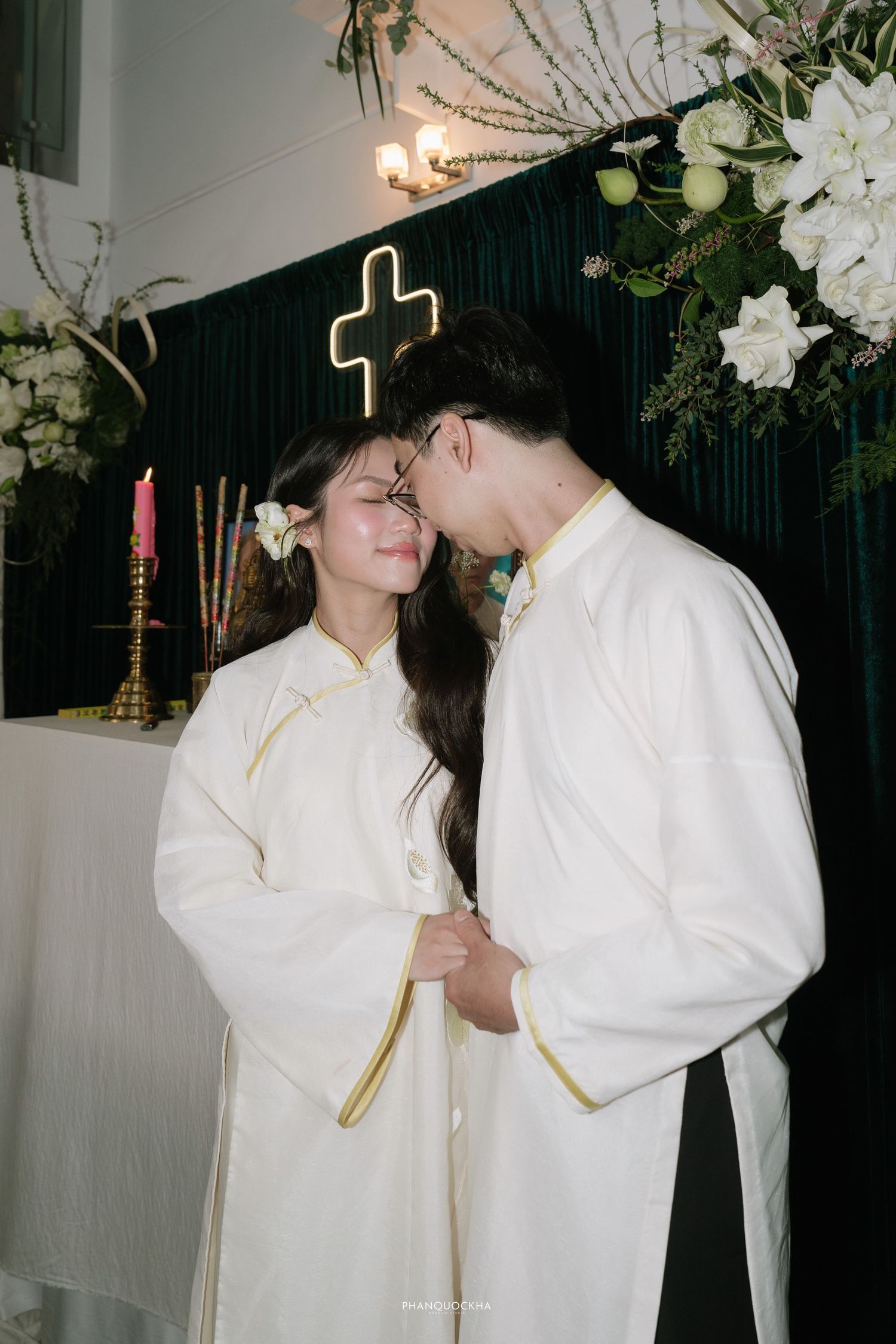 Vietnamese bride and groom in matching white ao dai touch foreheads tenderly before a cross altar.