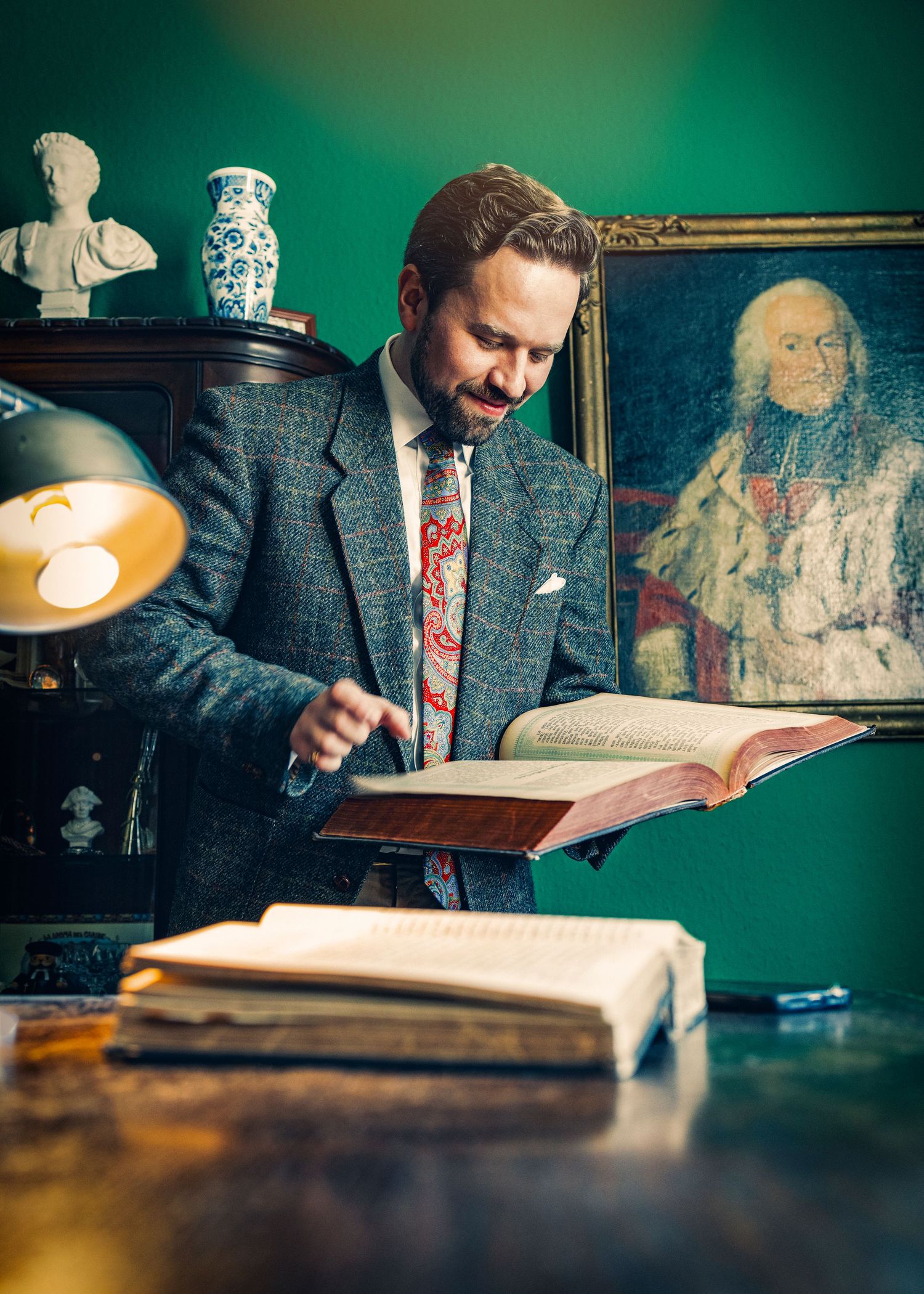 A person in a tweed jacket reads from a large antique book on a wooden desk in moody green lighting.
