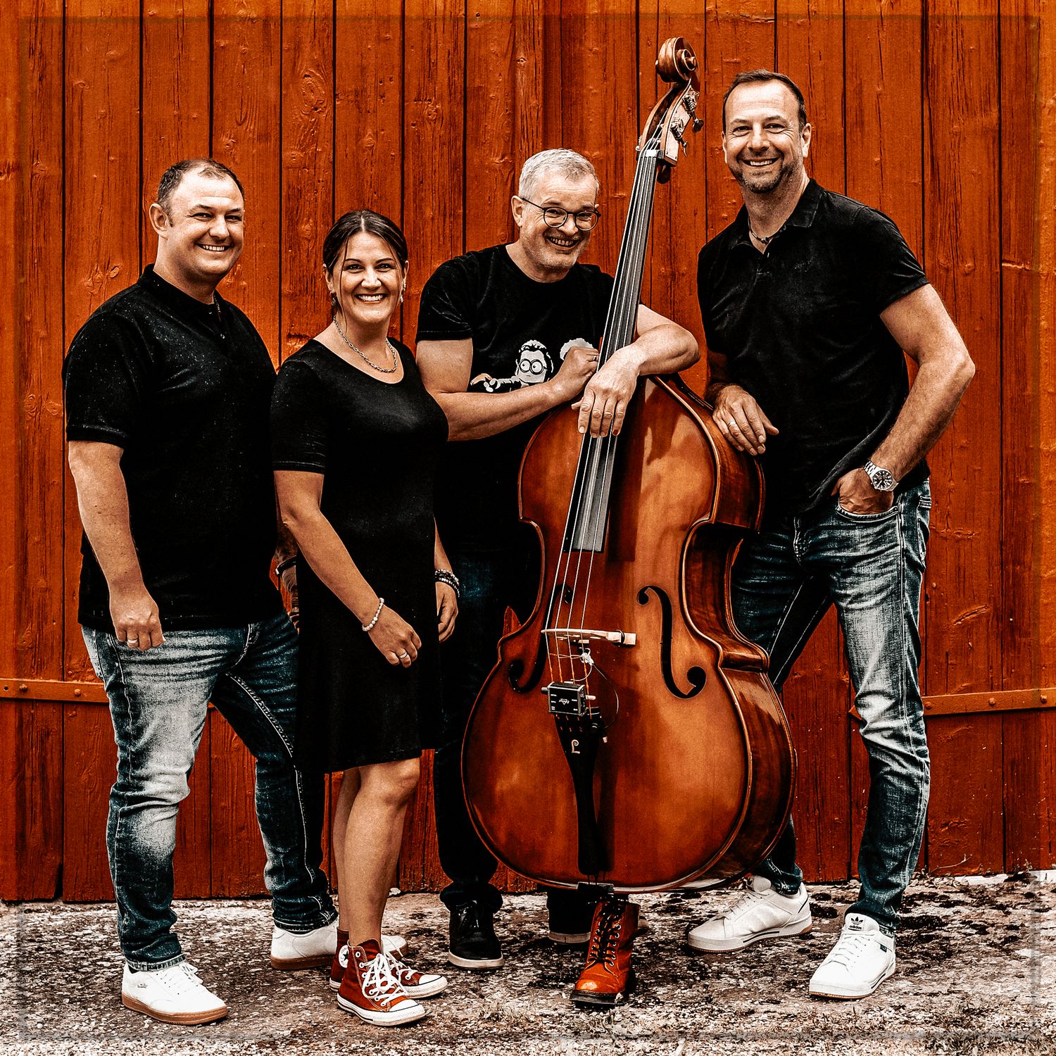 Musicians in black attire pose with instruments against a rustic red wooden wall background.