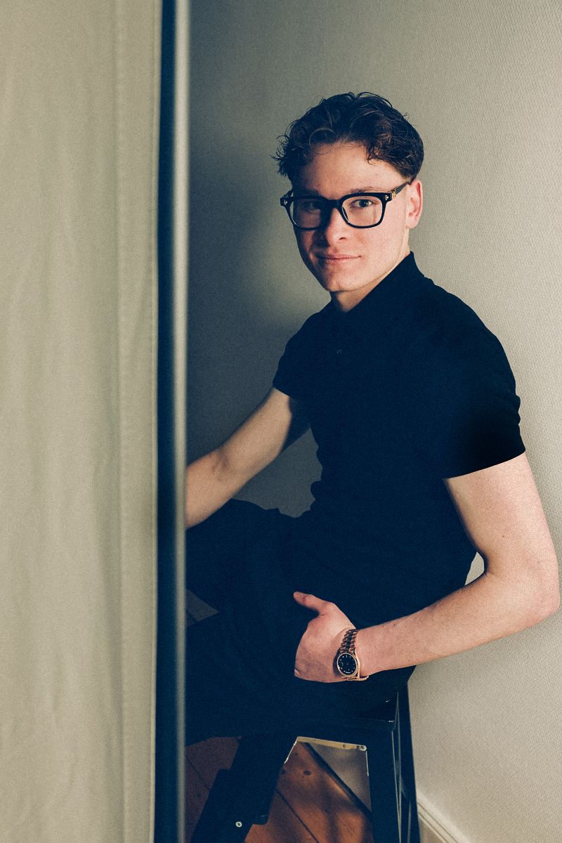 junger Mann mit stylischer schwarzer Brille und lässiger Haltung sitzend mit einer Hand in der Hosentasche im Fotostudio Aurelia Schulz