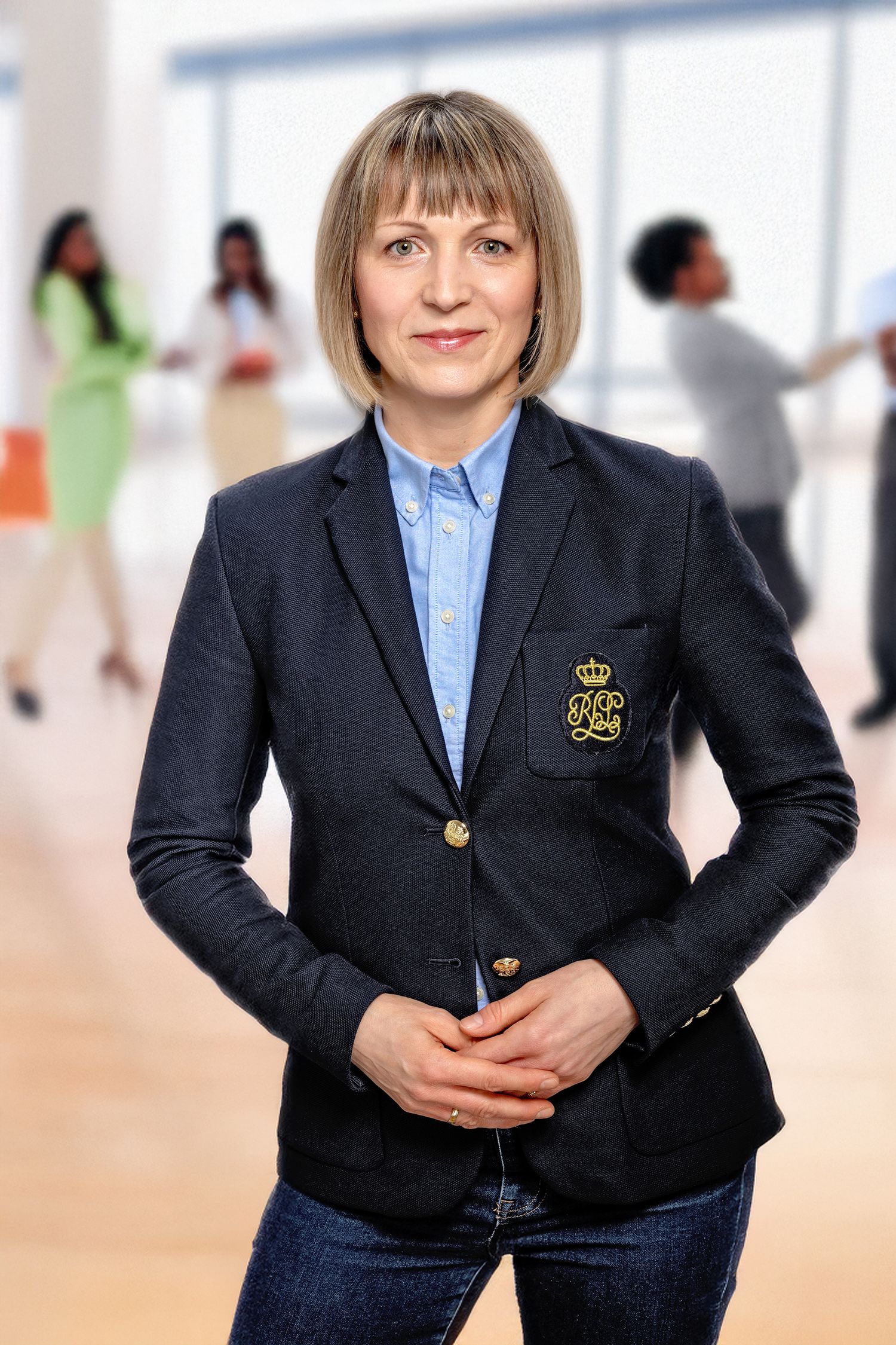 Professional woman in navy blazer with gold crest stands confidently in modern office with colleagues in background.