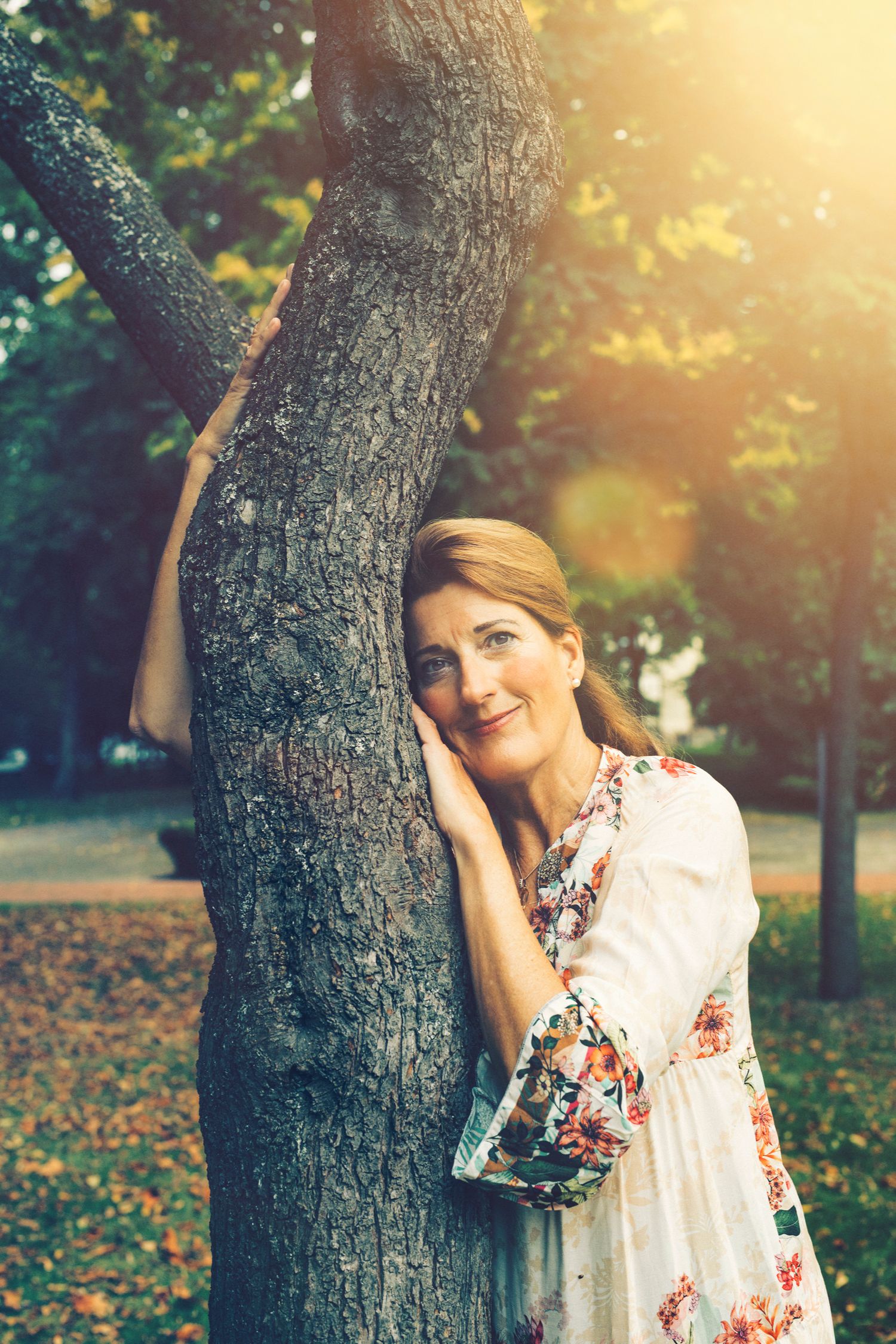 A person in a floral dress leans against a tree trunk in a sunlit autumn park with fallen leaves on the ground.