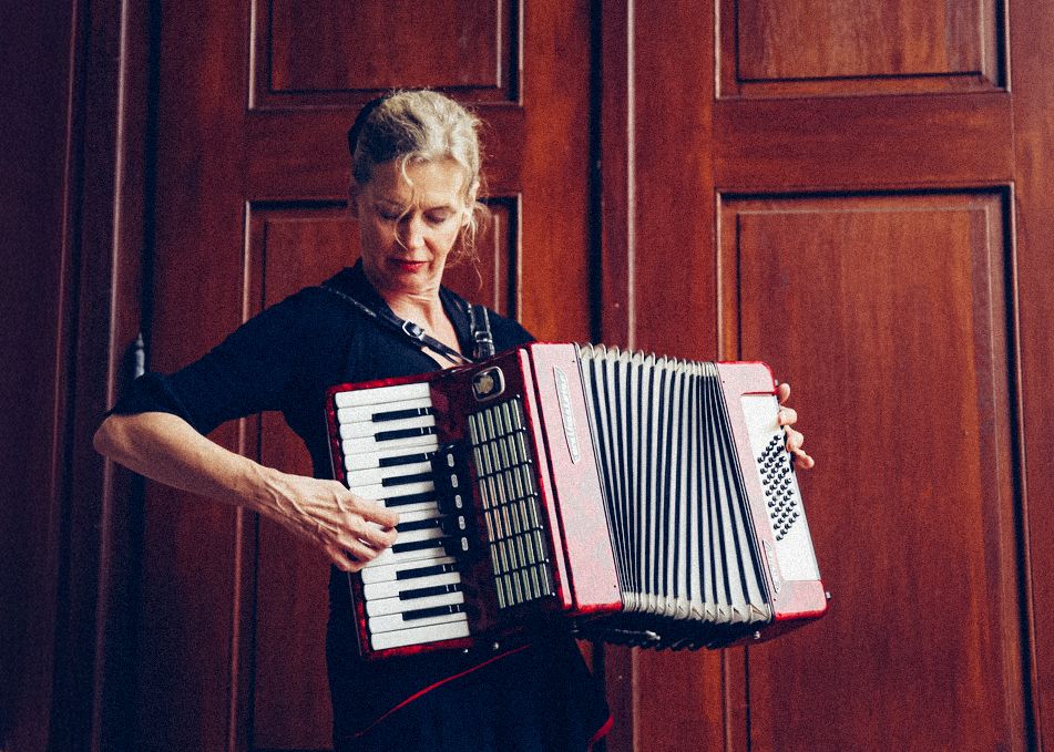 A musician performs with an accordion against a dark wooden paneled wall.