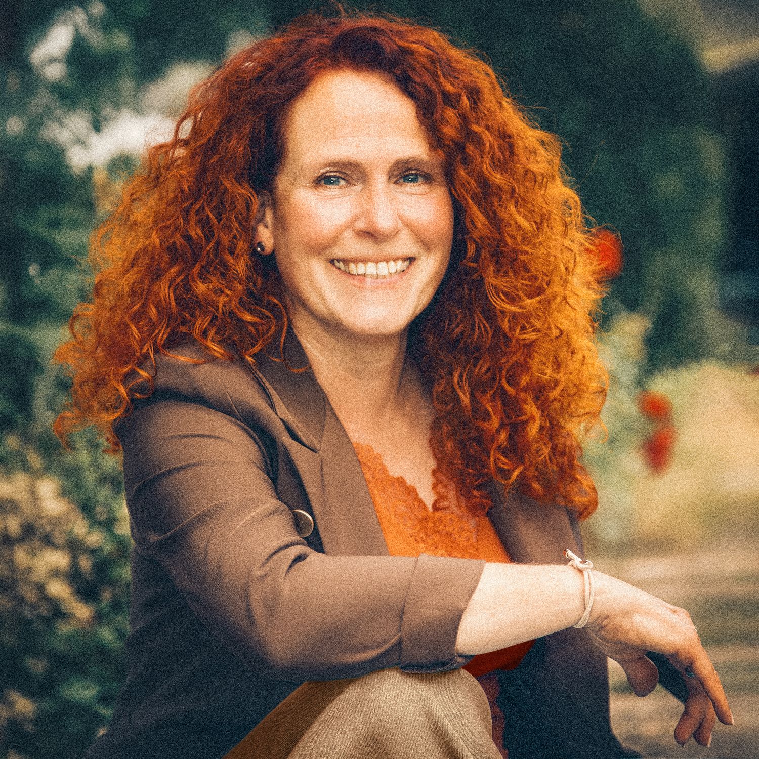 Woman with curly red hair wearing a brown blazer and orange top,  outdoors in a garden . Fröhliches lebendiges Outdoor Portrait einer Frau