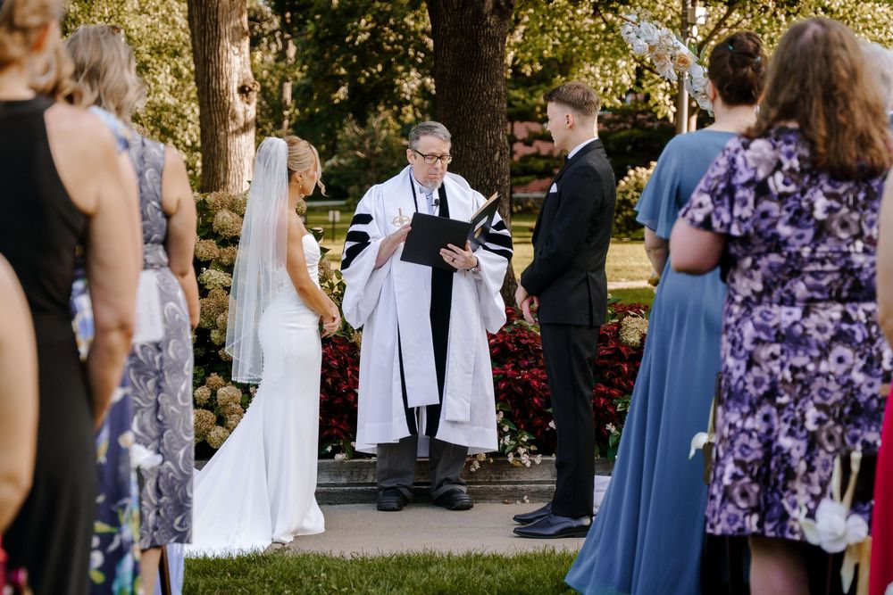 Outdoor wedding ceremony taking place in a garden setting with guests in blue dresses and officiant in white robes.