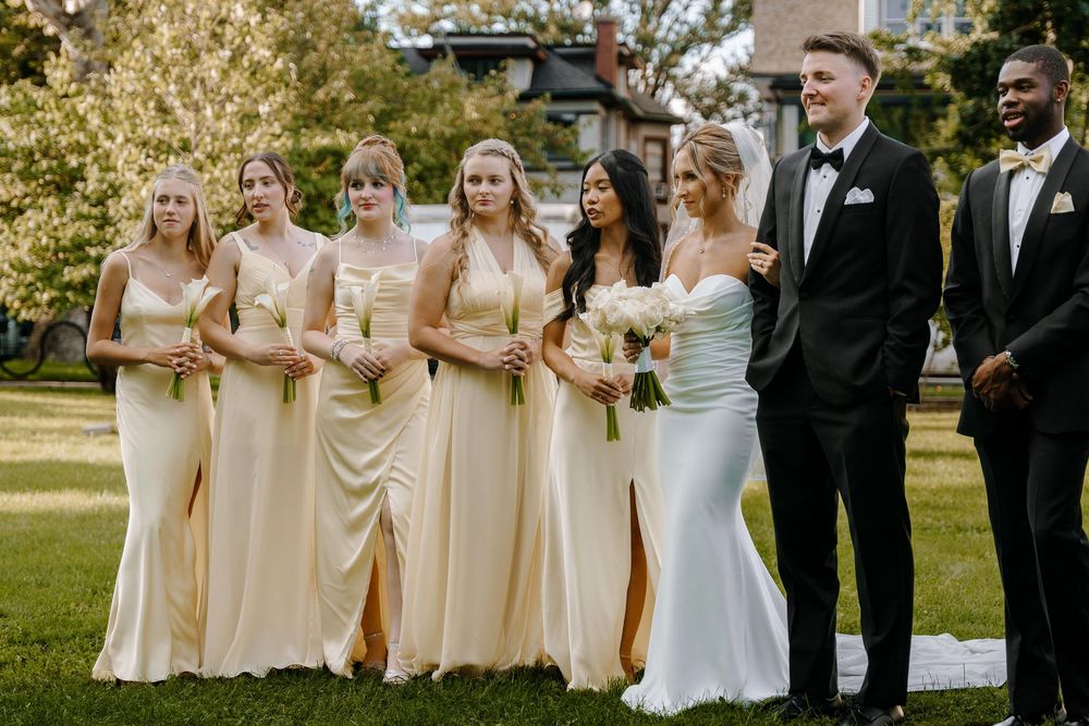 Wedding party in champagne-colored dresses and black suits posing outdoors on a sunny day.