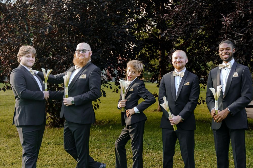 Groomsmen in black tuxedos laugh and smile together while standing in a line outdoors during a wedding celebration.