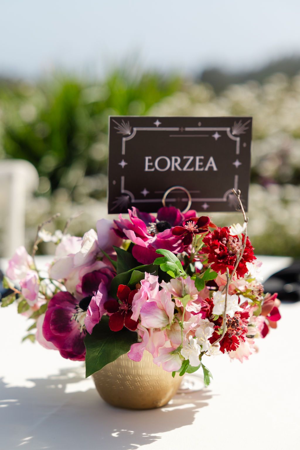 A colorful floral centerpiece with pink and burgundy flowers beside an Eorzea place card in a gold vase.