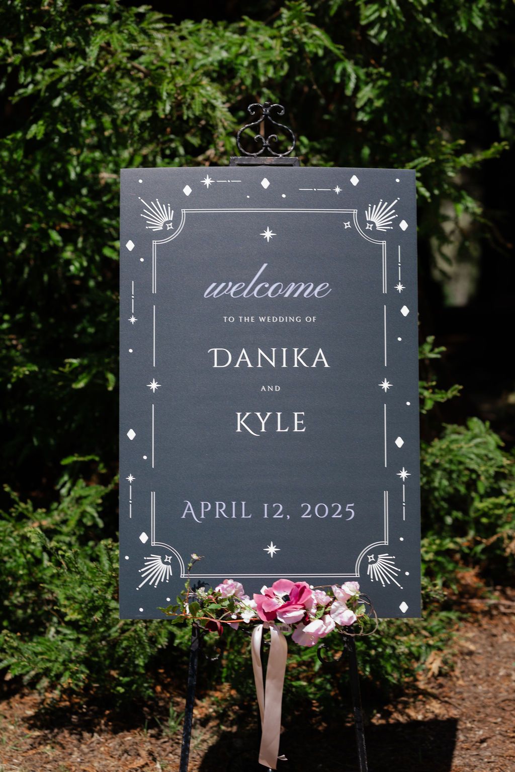 Pink and burgundy floral wedding arch stands elegantly against tall evergreen trees in an outdoor forest setting.