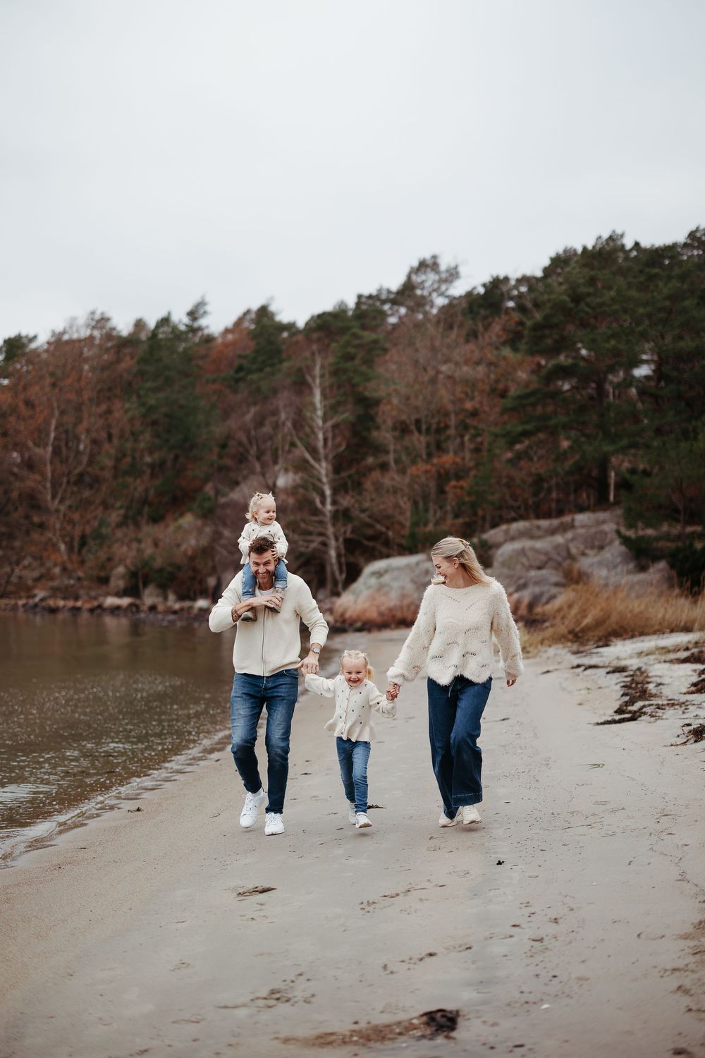 Family of four in cozy cream sweaters walks along a sandy beach, with a toddler on dad's shoulders and autumn trees behind.