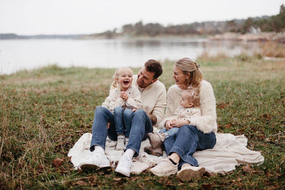 Parents and two young daughters sit laughing on a white blanket in a grassy field beside a calm lake in autumn.
