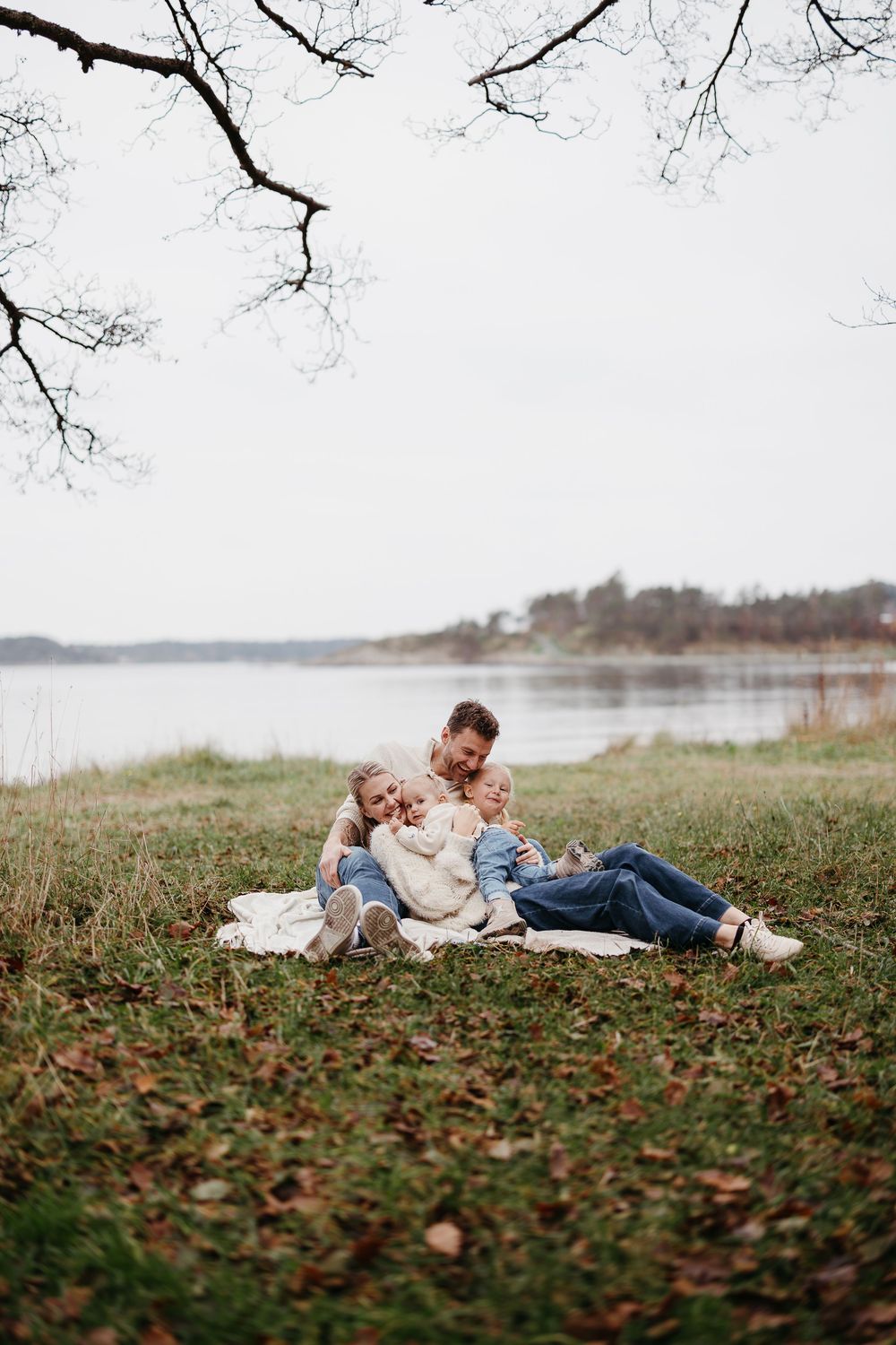 Family cuddles together on a blanket under bare tree branches on a grassy lakeside field during an overcast autumn day.