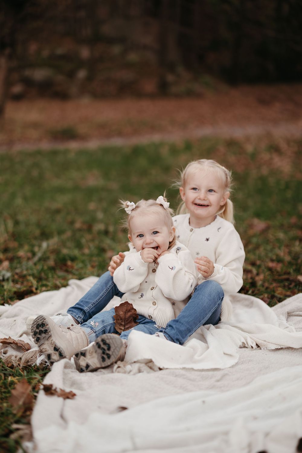Two young sisters in cream cardigans sit smiling on a white blanket on the grass during an autumn outdoor photo shoot.