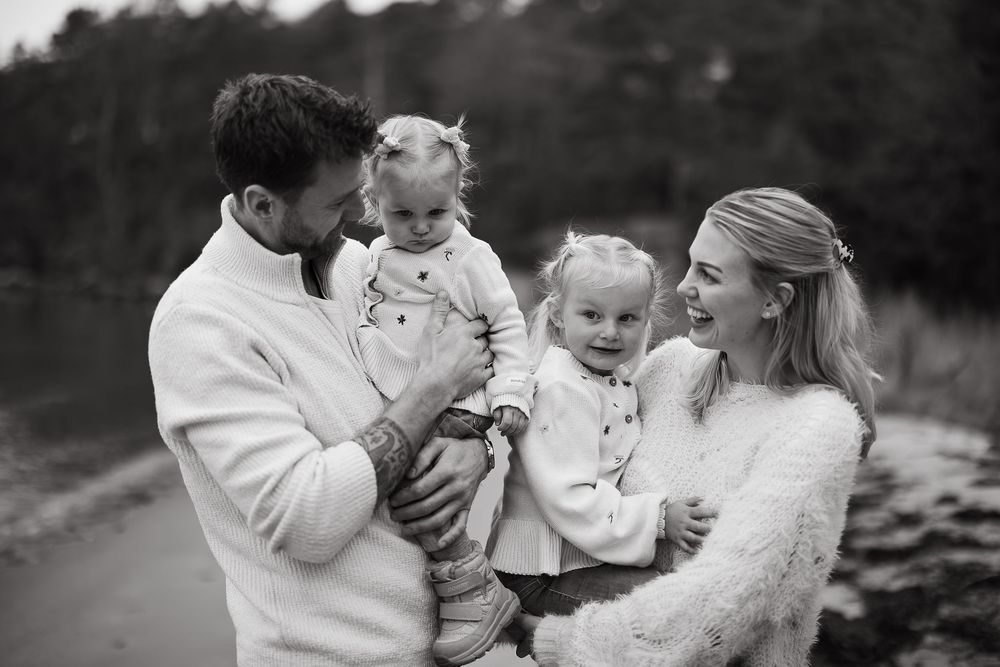 Black and white family portrait of two parents smiling while holding their two toddler daughters outdoors by the water.