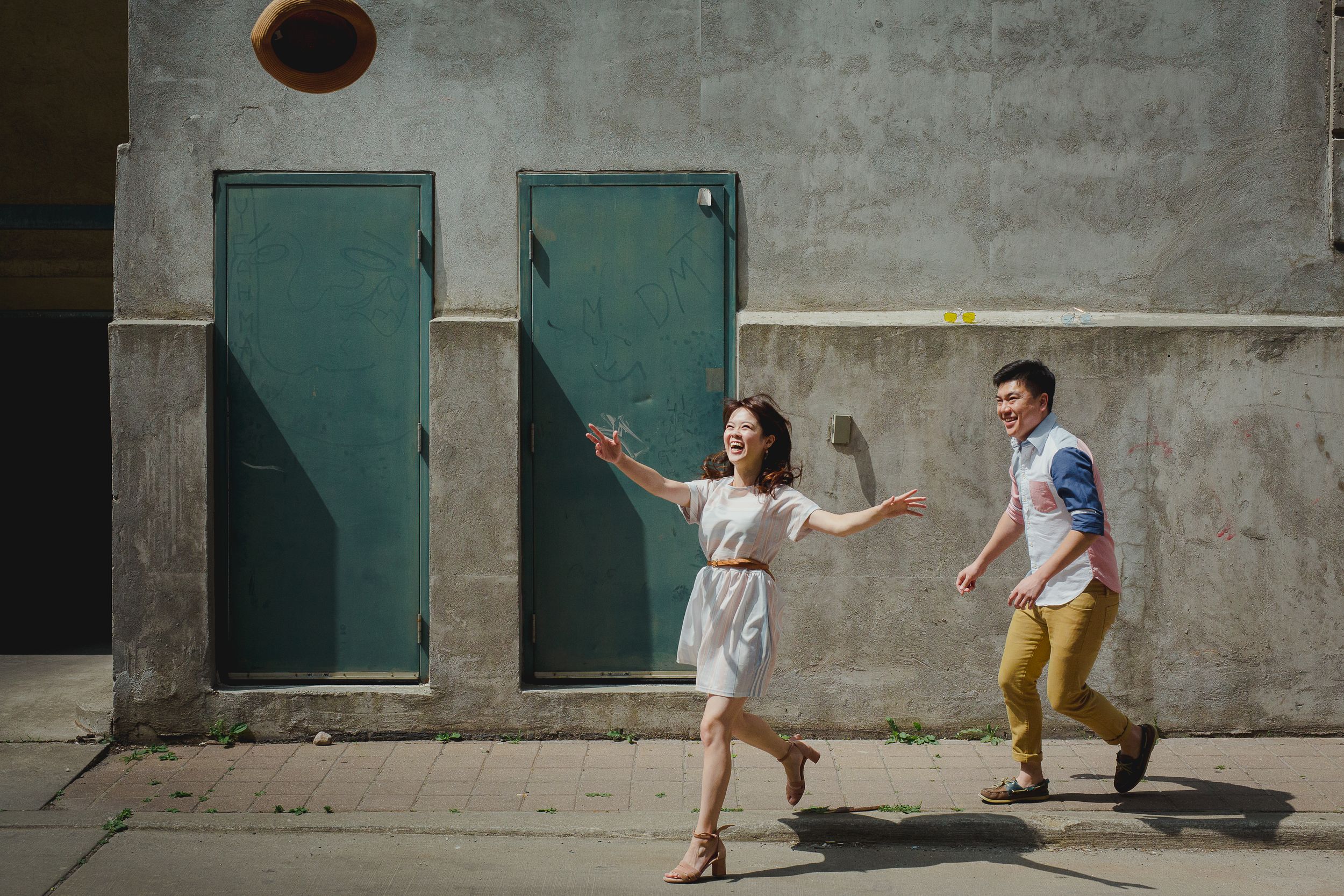 A couple in casual attire playfully runs and dances along a concrete wall in an urban setting.